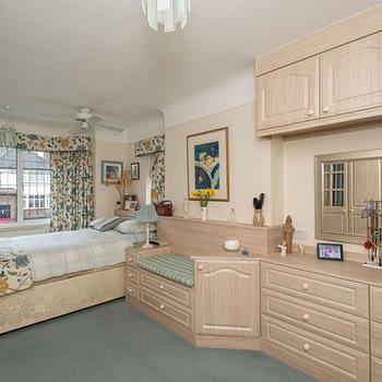 Bespoke fitted bedroom with angled cupboard, drawers, padded seat and bridging unit above, colour light swiss elm, style Athena